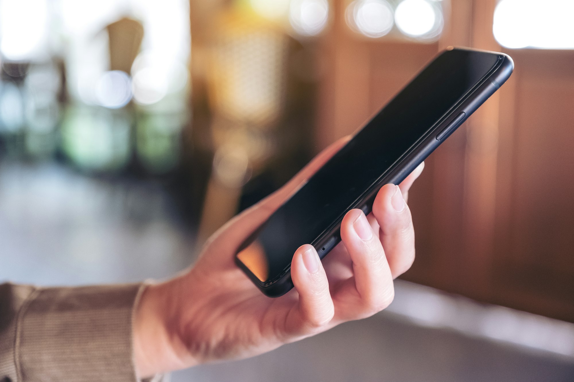 A woman holding and using a black smart phone with blur background in cafe